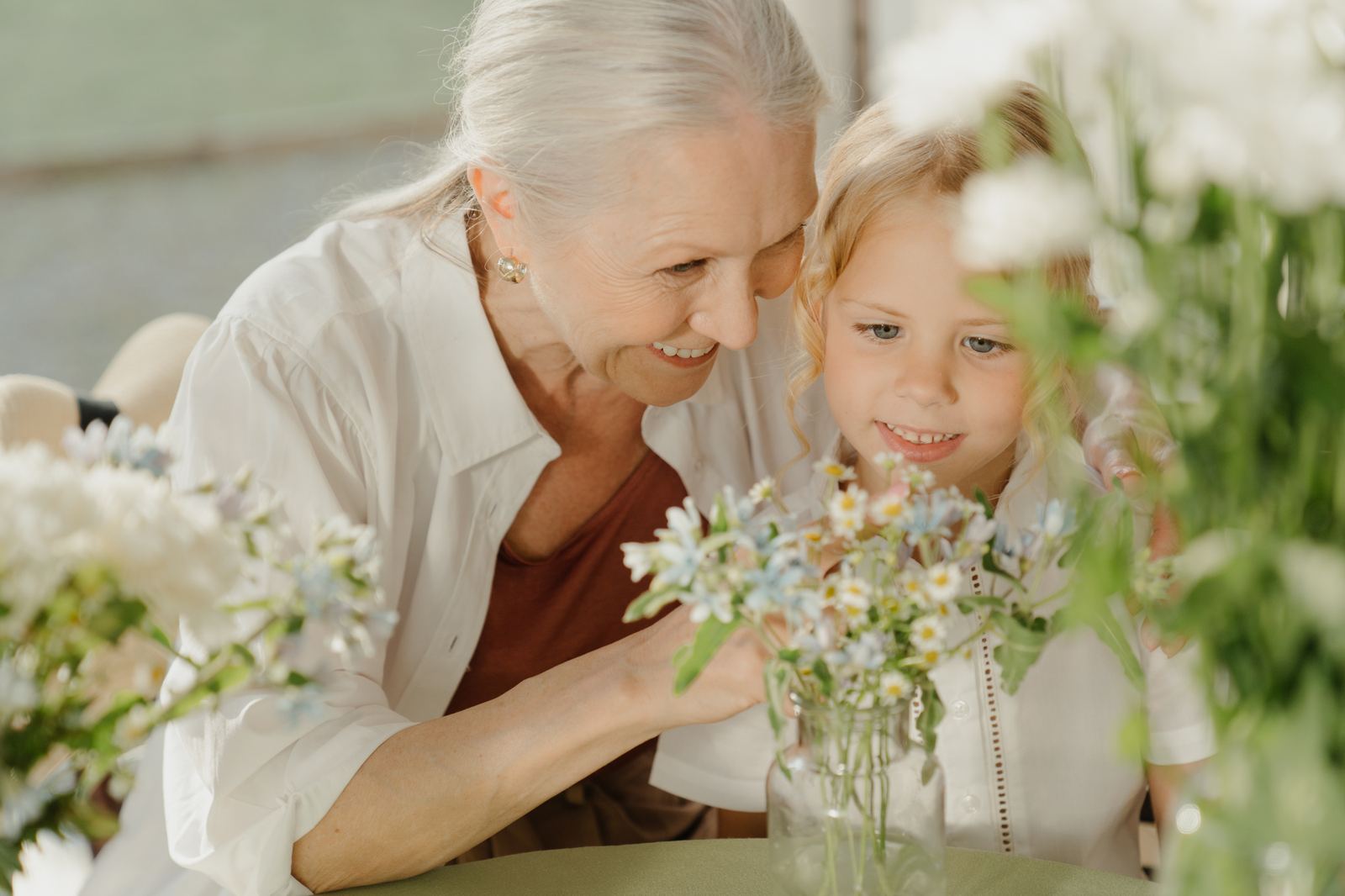 A grandmother and her granddaughter smiling together over a jar of wildflowers