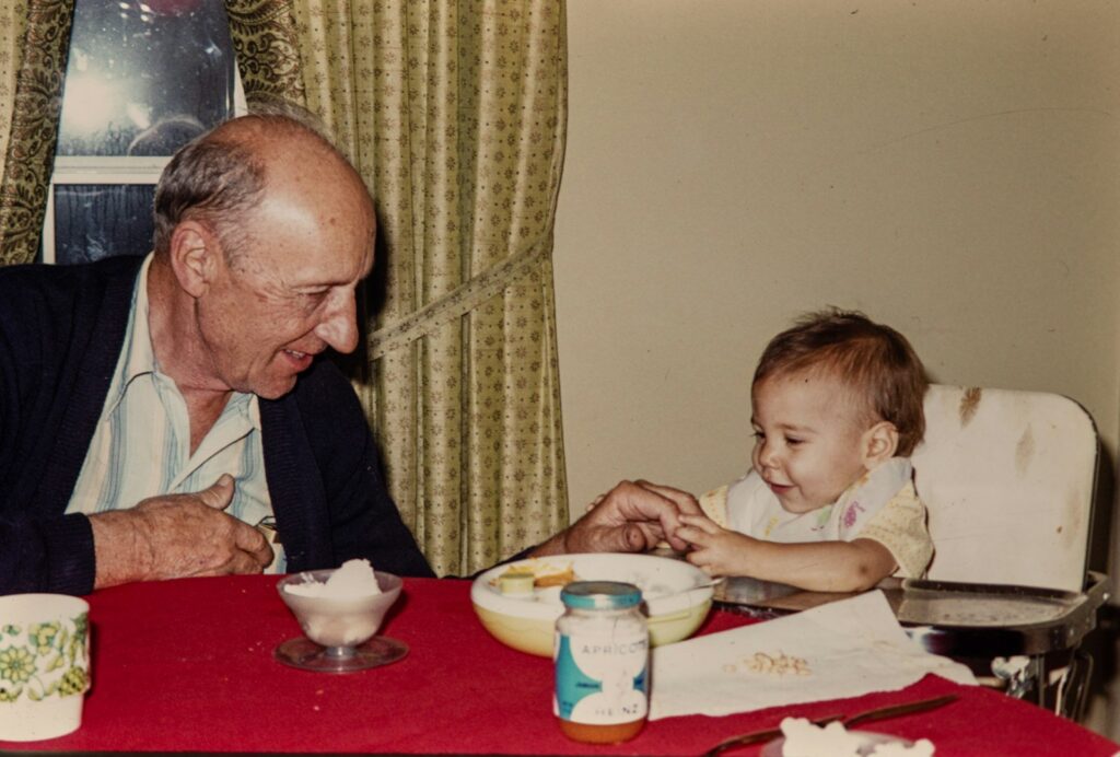 A 1980s snapshot of a grandfather feeding a baby at the table